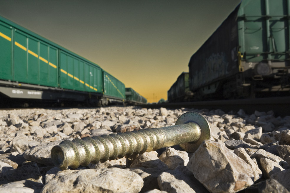 En primer plano un tirafondo centrado, sobre la piedra del balasto. A los lados dos trenes de mercancías que confluyen en el horizonte.